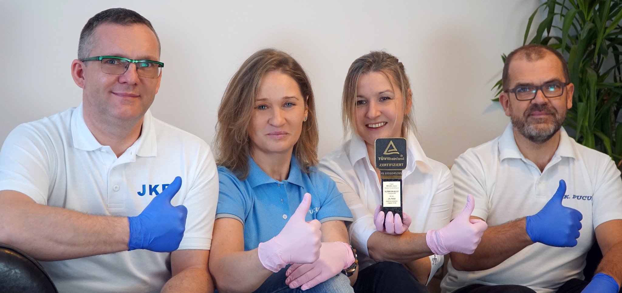 Zespół w rękawicach prezentuje certyfikat TÜV Rheinland, kciuki w górę. W tle roślina doniczkowa i biała ściana. Profesjonalizm i jakość usług.