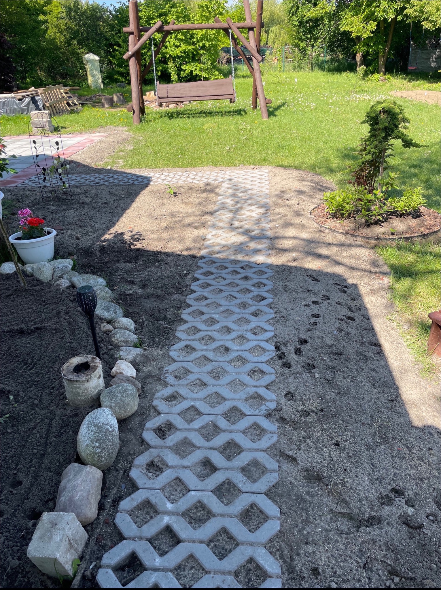 Ścieżka z ażurowych płyt betonowych prowadząca przez ogród, z drewnianą huśtawką w tle. Elementy dekoracyjne z kamieni i roślin. Ślady na ziemi.