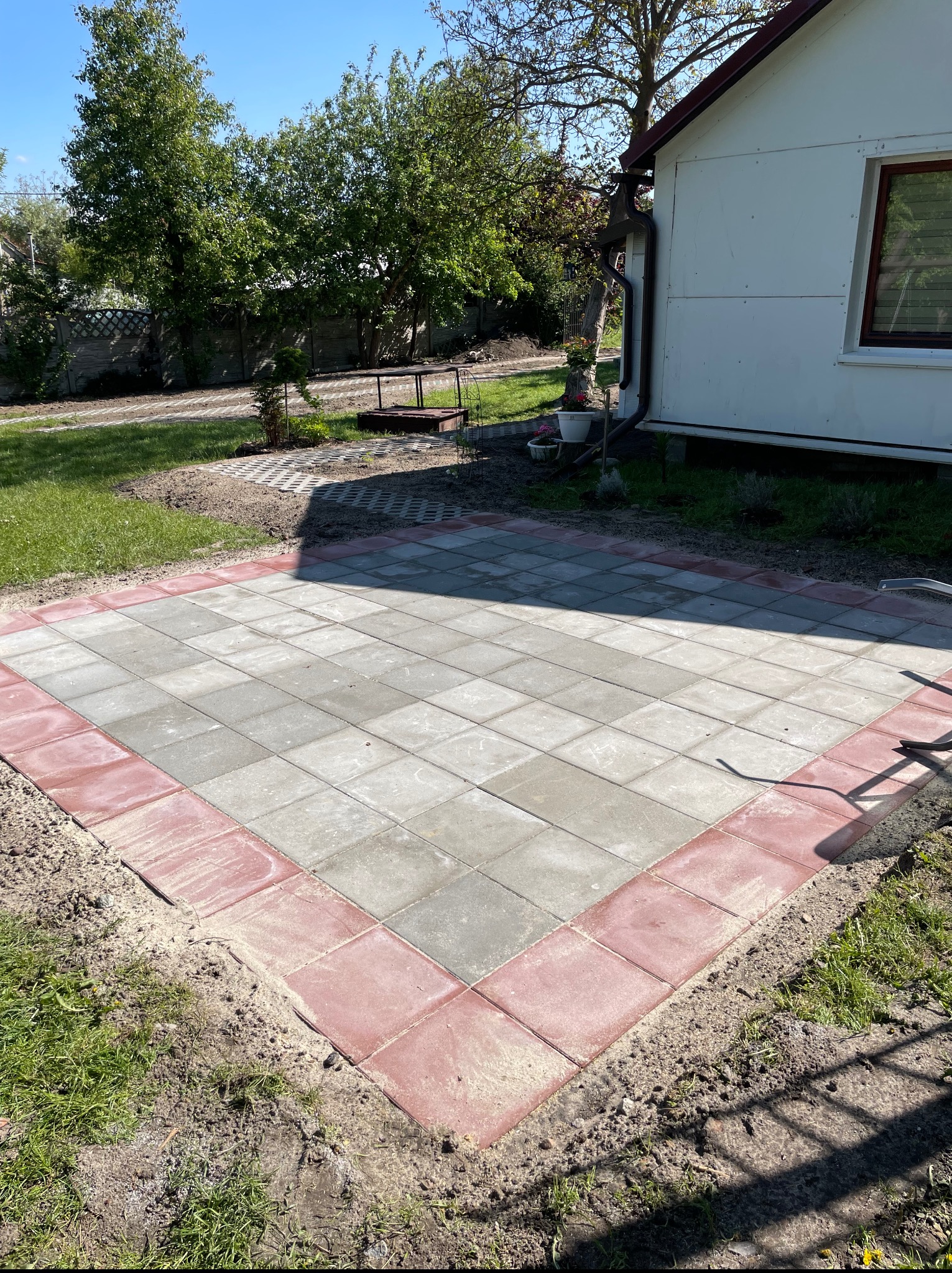 Ułożona kostka brukowa w szaro-czerwonej kolorystyce tworząca patio przy białym budynku, z widocznym trawnikiem i drzewami w tle, w słoneczny dzień.