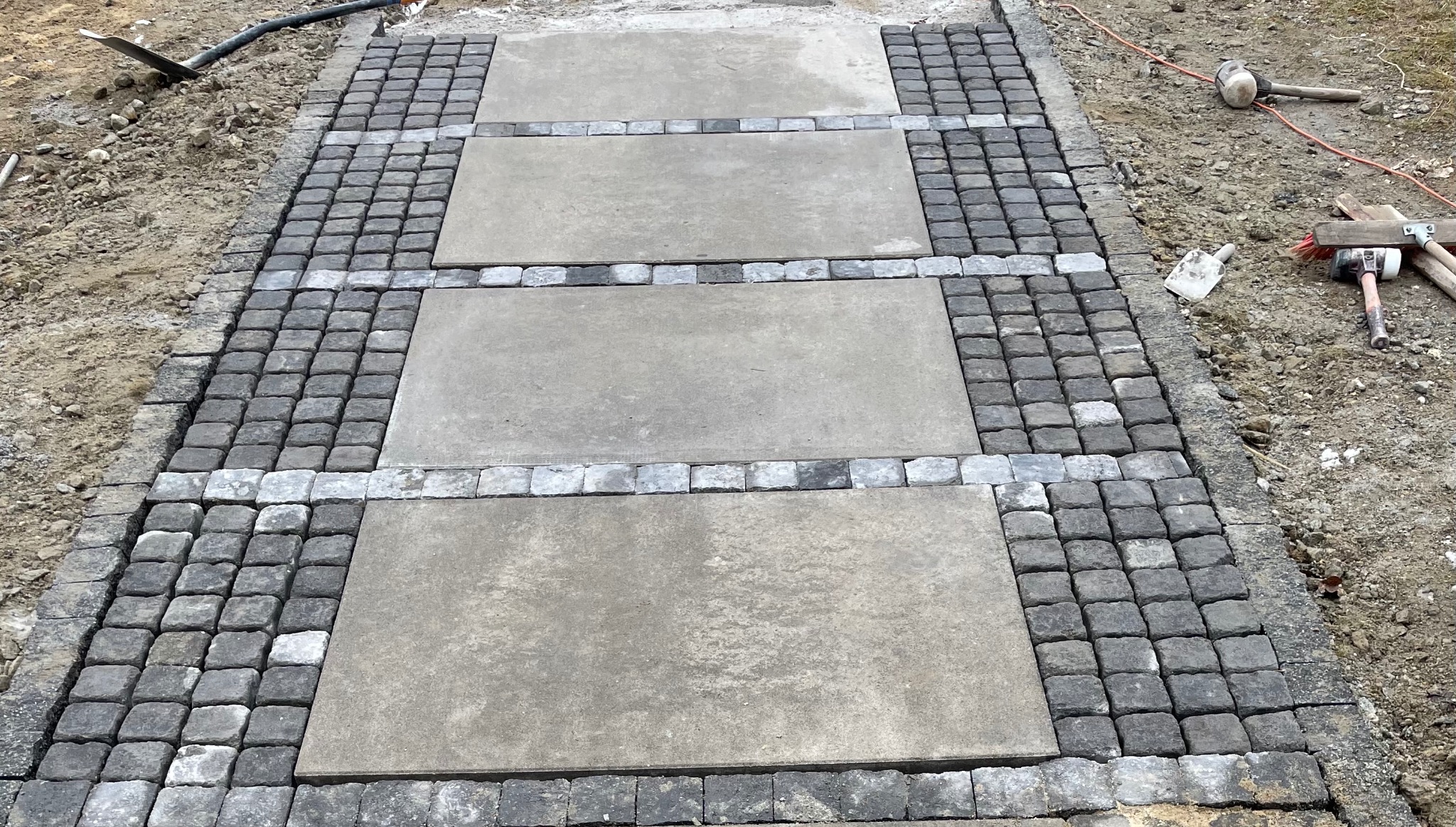 Ścieżka z dużych, szarych płyt betonowych, obramowana ciemną kostką brukową i jaśniejszymi kostkami, widok z góry, narzędzia pracy widoczne w tle.