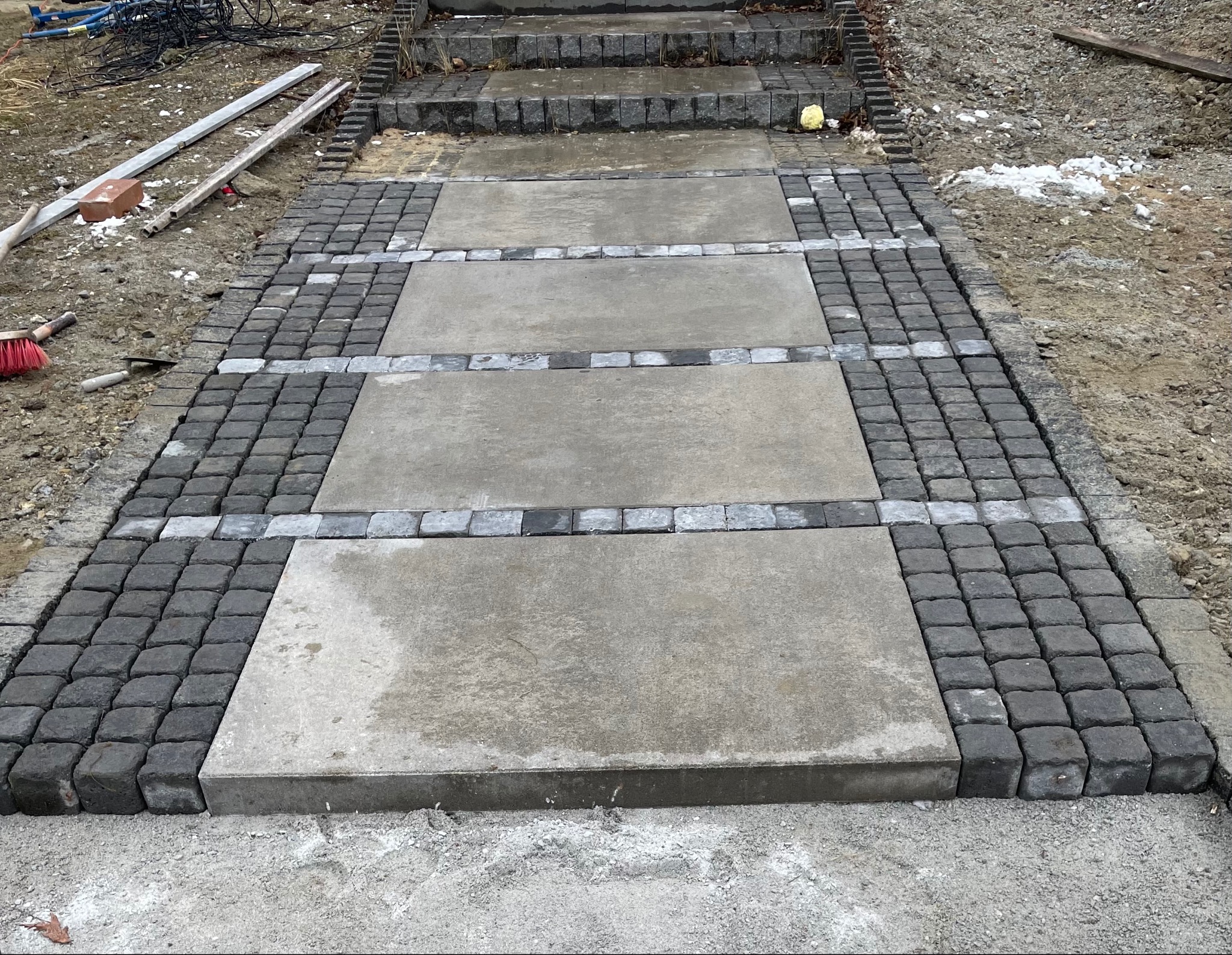 Ścieżka z betonowych płyt obłożona ciemną kostką brukową, prowadząca do schodów. Widoczne narzędzia i resztki materiałów budowlanych. Kąt ujęcia z dołu.