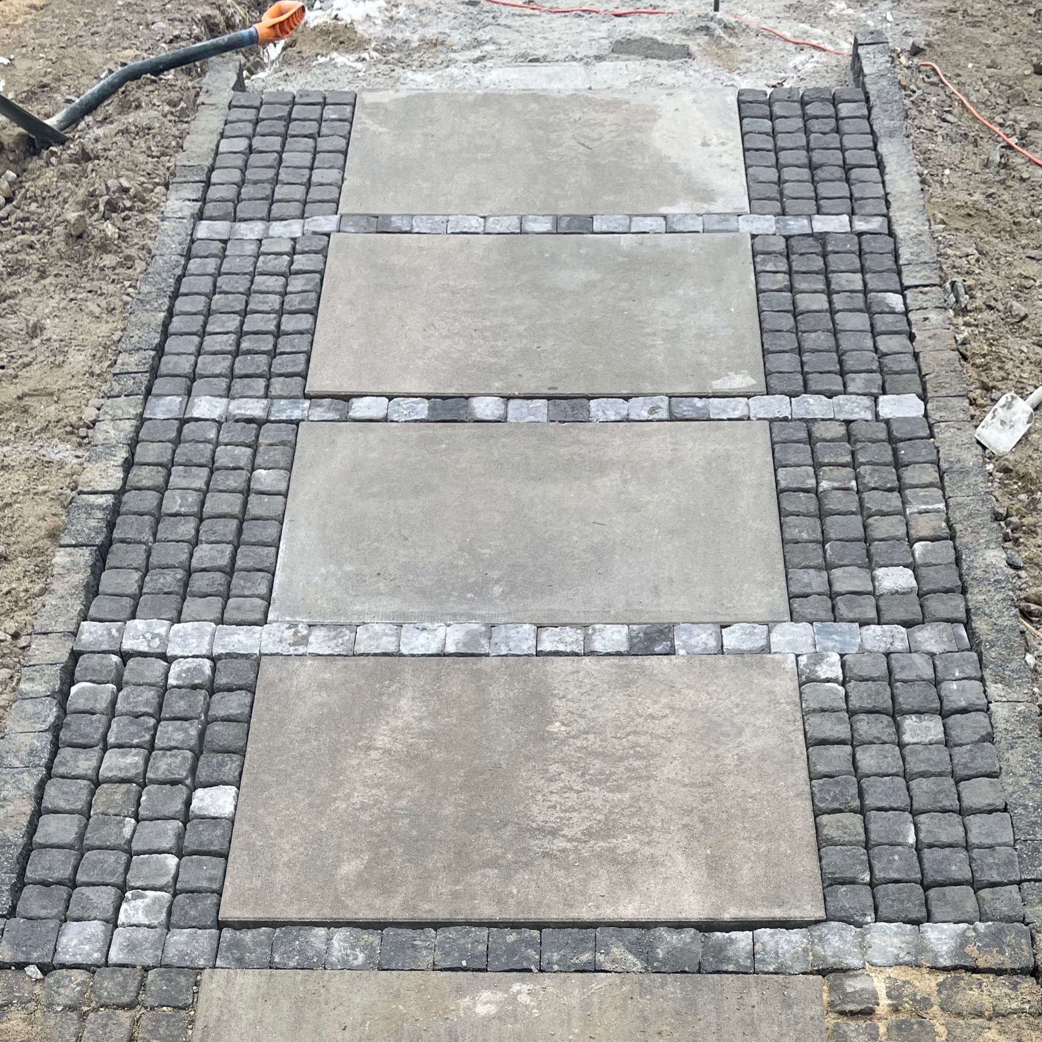 Ścieżka z szarej kostki brukowej i betonowych płyt, otoczona ziemią. Widoczne narzędzia i przewody. Układanie kostki w trakcie realizacji.