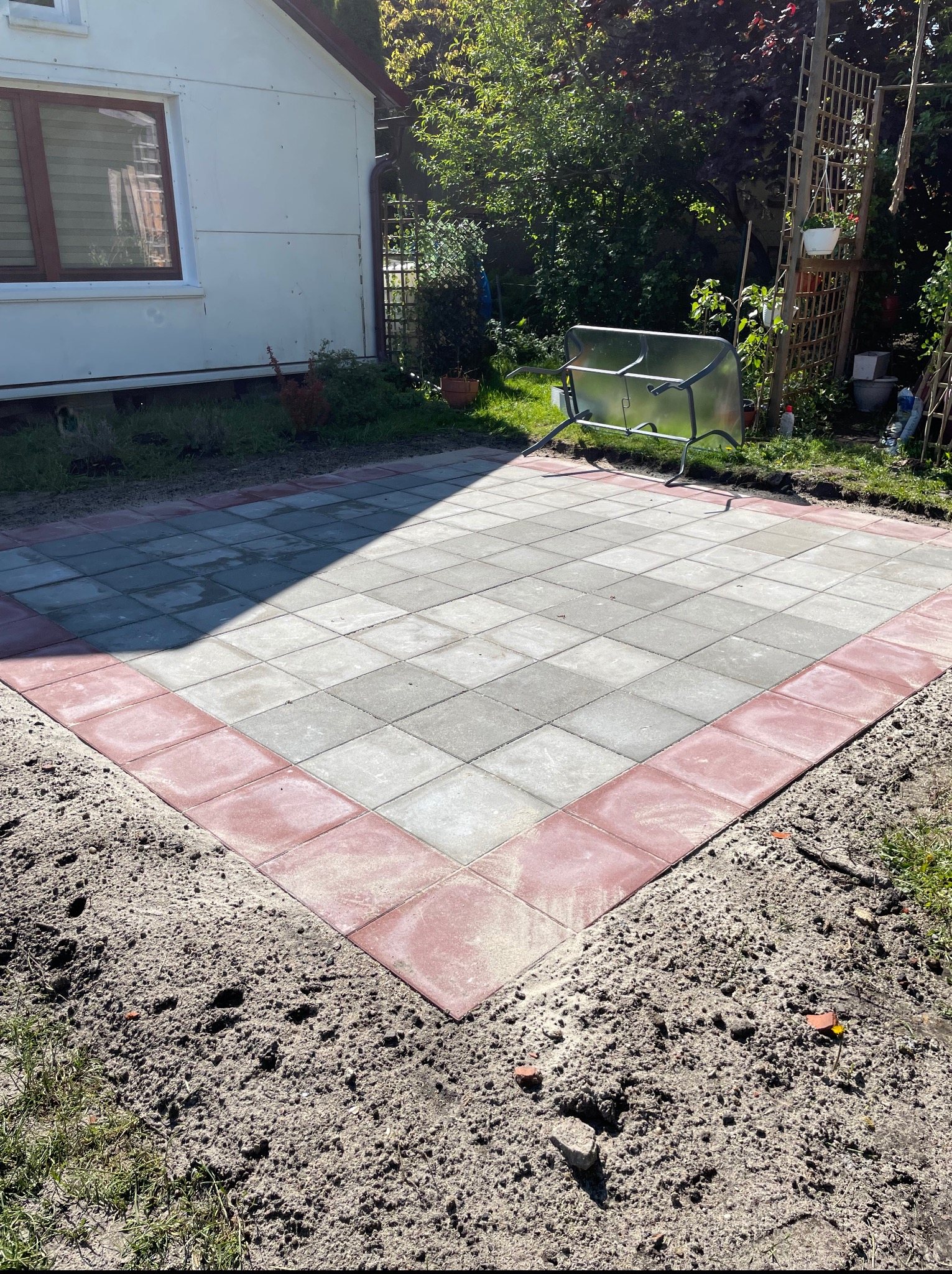 Ułożony plac z kostki brukowej w odcieniach szarości z bordową obwódką, widok z góry, na tle domu i zieleni. Widoczne nierówności terenu po bokach.