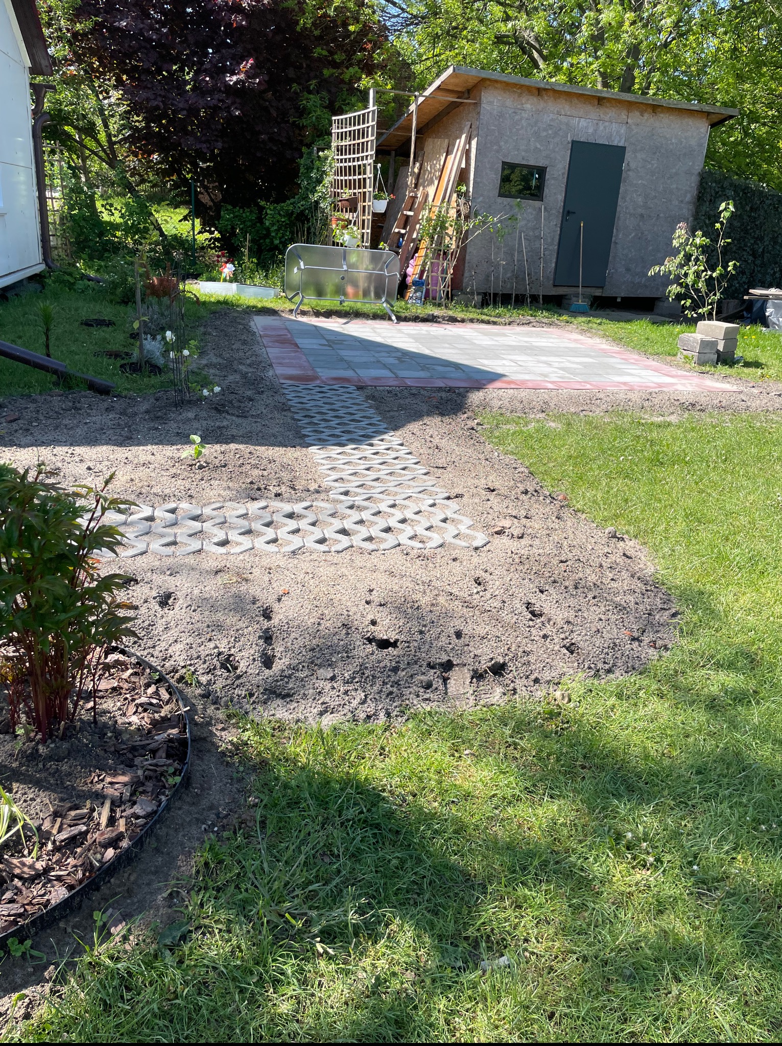 Ścieżka z ażurowych płyt prowadzi do ułożonej z kostki strefy relaksu w ogrodzie. Widoczny fragment trawnika, rabata i drewniany budynek gospodarczy w tle.