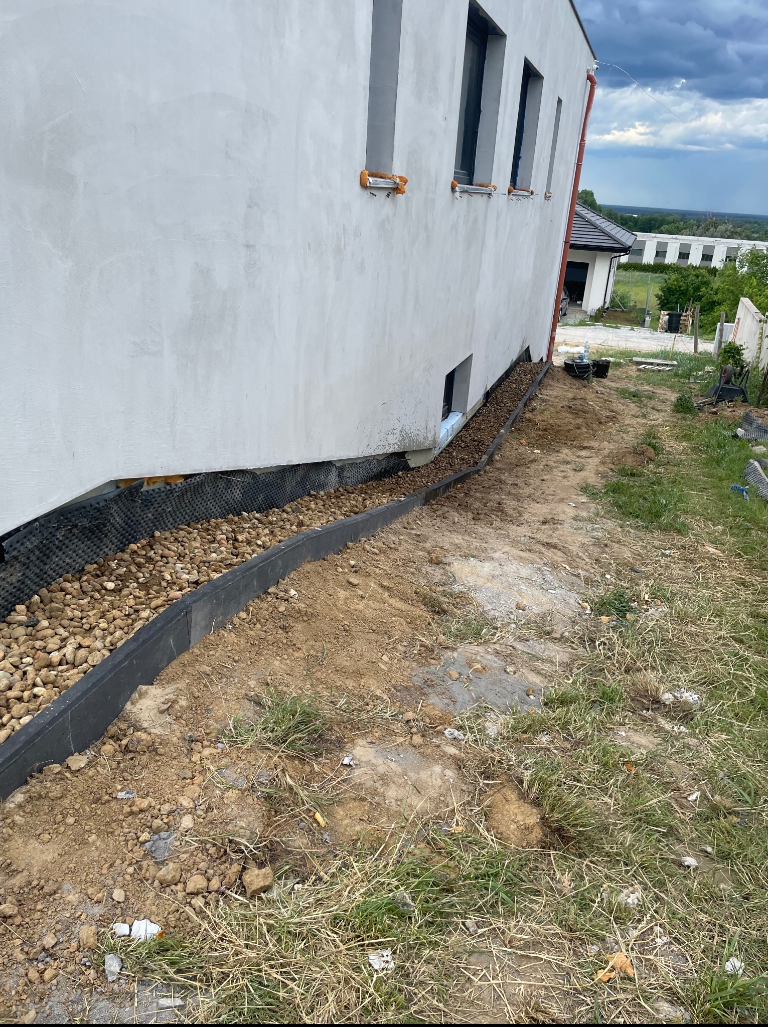 Drenaż opaskowy budynku w trakcie realizacji: czarna listwa obrzeżna, kamienie drenażowe, folia kubełkowa. Budynek w trakcie wykończenia, widok na otoczenie.