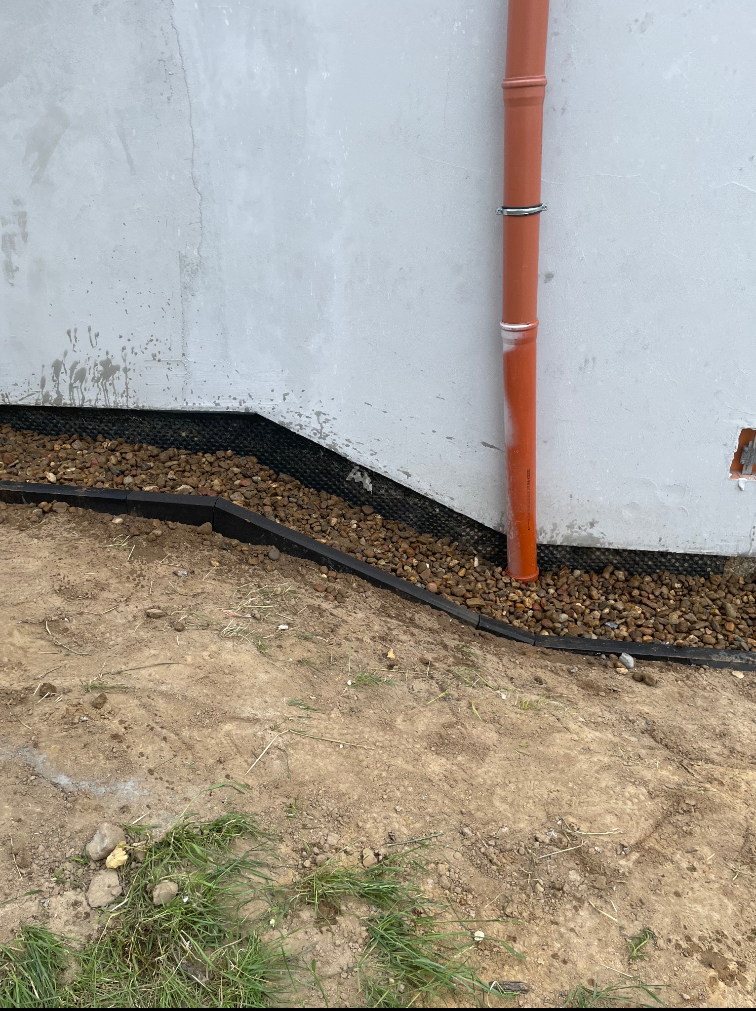 Fundament budynku z izolacją, drenażem z kamyków i rurą spustową. Widoczne elementy instalacji i warstwy izolacyjne.