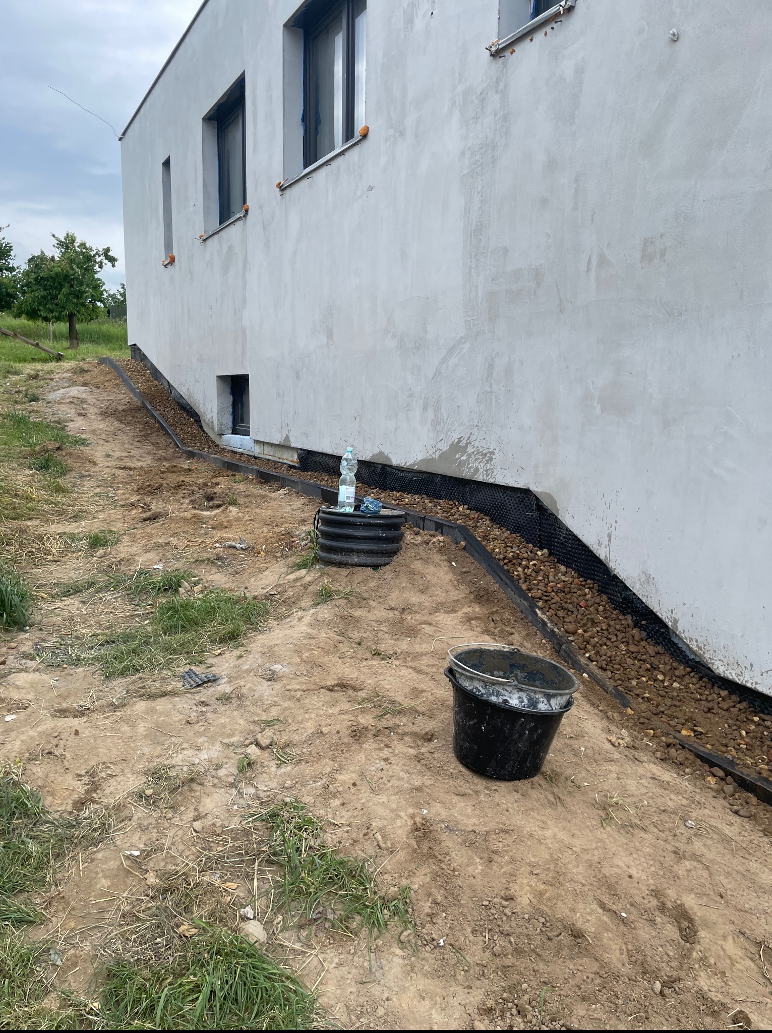 Przyścienny drenaż opaskowy z folią kubełkową i kamieniami, biegnący wzdłuż fundamentu jasnego budynku. Widoczne okna piwniczne i wiadro budowlane.