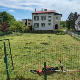 Trawa skoszona na posesji, z kosiarką spalinową przy zielonym ogrodzeniu, przed domem z czerwonym dachem i błękitnym niebem w tle.