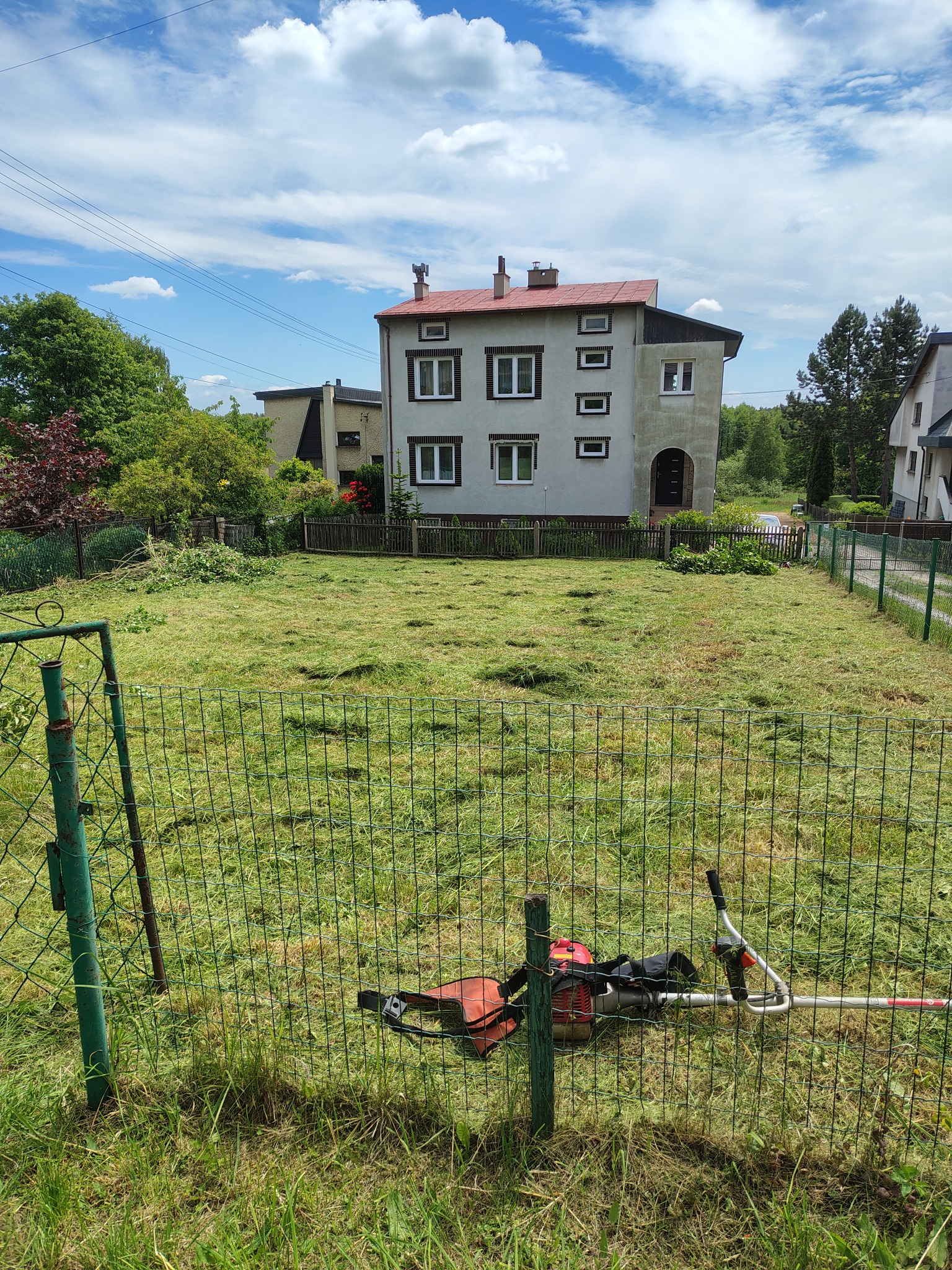 Trawa skoszona na posesji, z kosiarką spalinową przy zielonym ogrodzeniu, przed domem z czerwonym dachem i błękitnym niebem w tle.