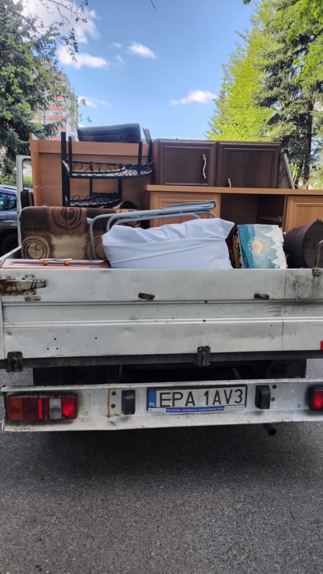 Tył białej ciężarówki wypełnionej meblami i przedmiotami gospodarstwa domowego, widoczne tablice rejestracyjne. Drzewa w tle sugerują miejską lokalizację.