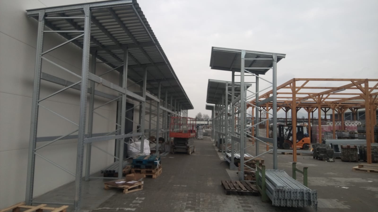 Konstrukcja stalowa zadaszenia magazynowego w trakcie budowy. Widoczne elementy konstrukcyjne, palety z materiałami i wózek widłowy w tle.