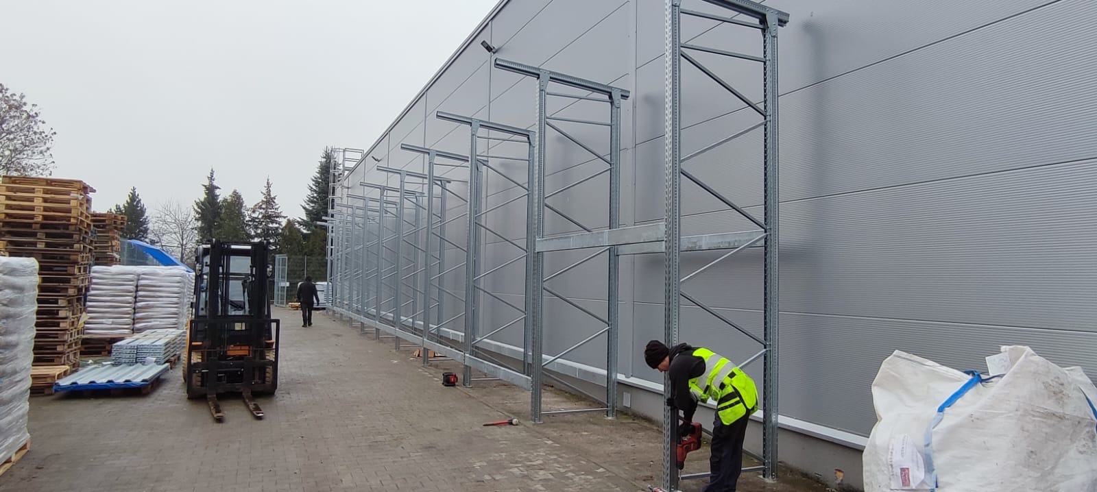 Montaż regałów magazynowych przy hali. Pracownik w kamizelce wierci otwory w metalowej konstrukcji. W tle wózek widłowy i materiały budowlane.