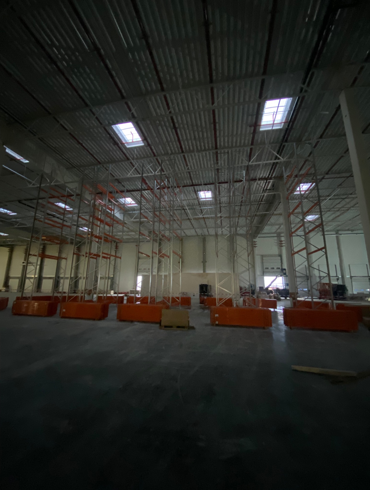 Wnętrze hali w trakcie budowy z widocznymi elementami konstrukcji regałów magazynowych i materiałami składowanymi na podłodze. Widoczne świetliki w dachu hali.