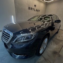 Autokosmetyka Brylant - Błyszczący, czarny Mercedes S-klasa po detailingu, widoczny z boku na tle szarej ściany z logo 'Brylant' i heksagonalnym oświetleniem sufitowym.