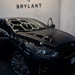 Błyszczący, czarny samochód Kia z otwartymi drzwiami, odbijający światła w kształcie plastra miodu. Logo 'Brylant' na ścianie w tle. Auto po detailingu.
