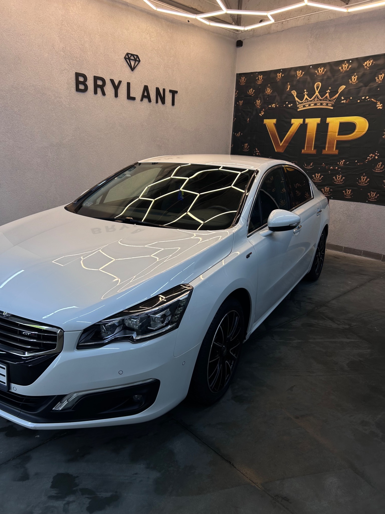 Biały samochód marki Peugeot w myjni, z logo 'Brylant' i banerem 'VIP' w tle, oświetlony ledowym światłem w kształcie plastra miodu.