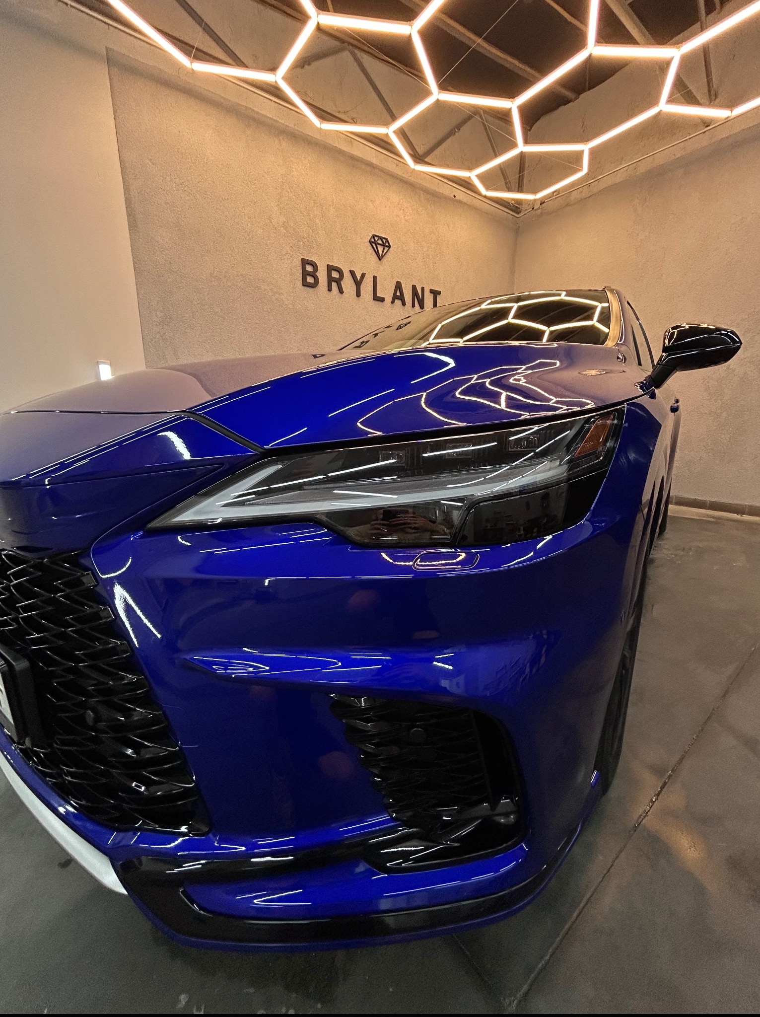 Błyszczący, niebieski Lexus RX po detailingu, widoczny z bliska, z odbiciami światła na lakierze i logiem 'Brylant' w tle, pod nowoczesnym oświetleniem.