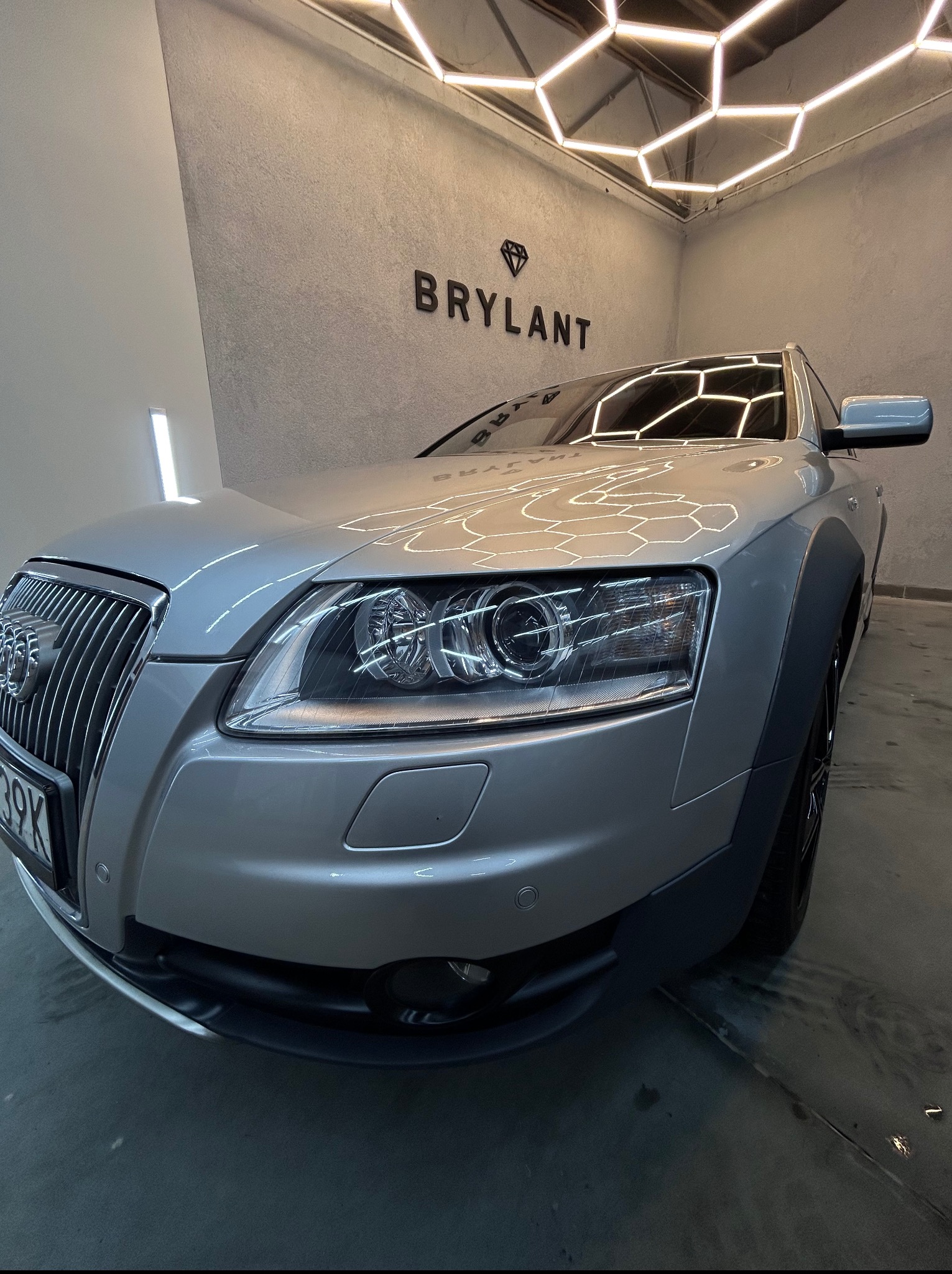 Błyszczący, srebrny Audi Allroad w myjni z heksagonalnym oświetleniem sufitowym. Widoczne logo 'Brylant' na ścianie. Auto po detailingu, z bliska, reflektory.