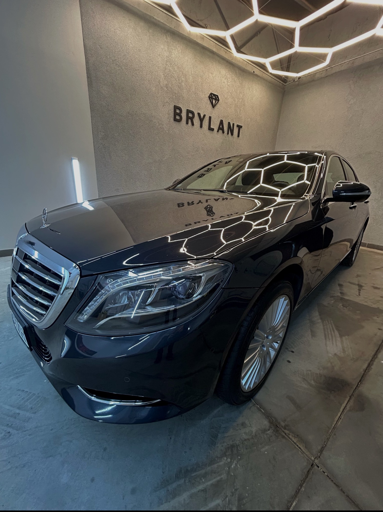 Błyszczący, czarny Mercedes S-klasa po detailingu, widoczny z boku na tle szarej ściany z logo 'Brylant' i heksagonalnym oświetleniem sufitowym.