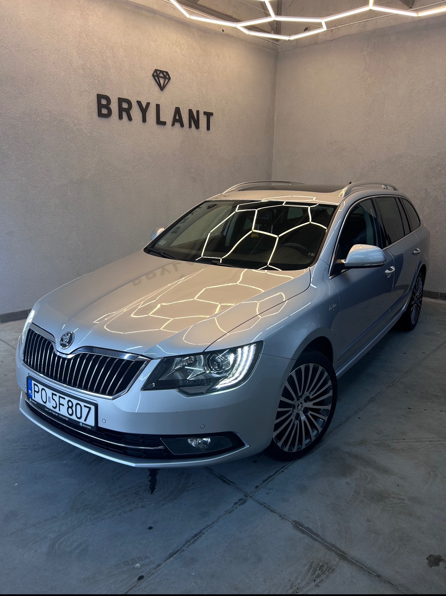 Srebrna Skoda Superb Combi po zabiegach w myjni 'Brylant', z widocznym logo firmy. Oświetlenie sufitowe odbija się na lakierze, podkreślając jego czystość.