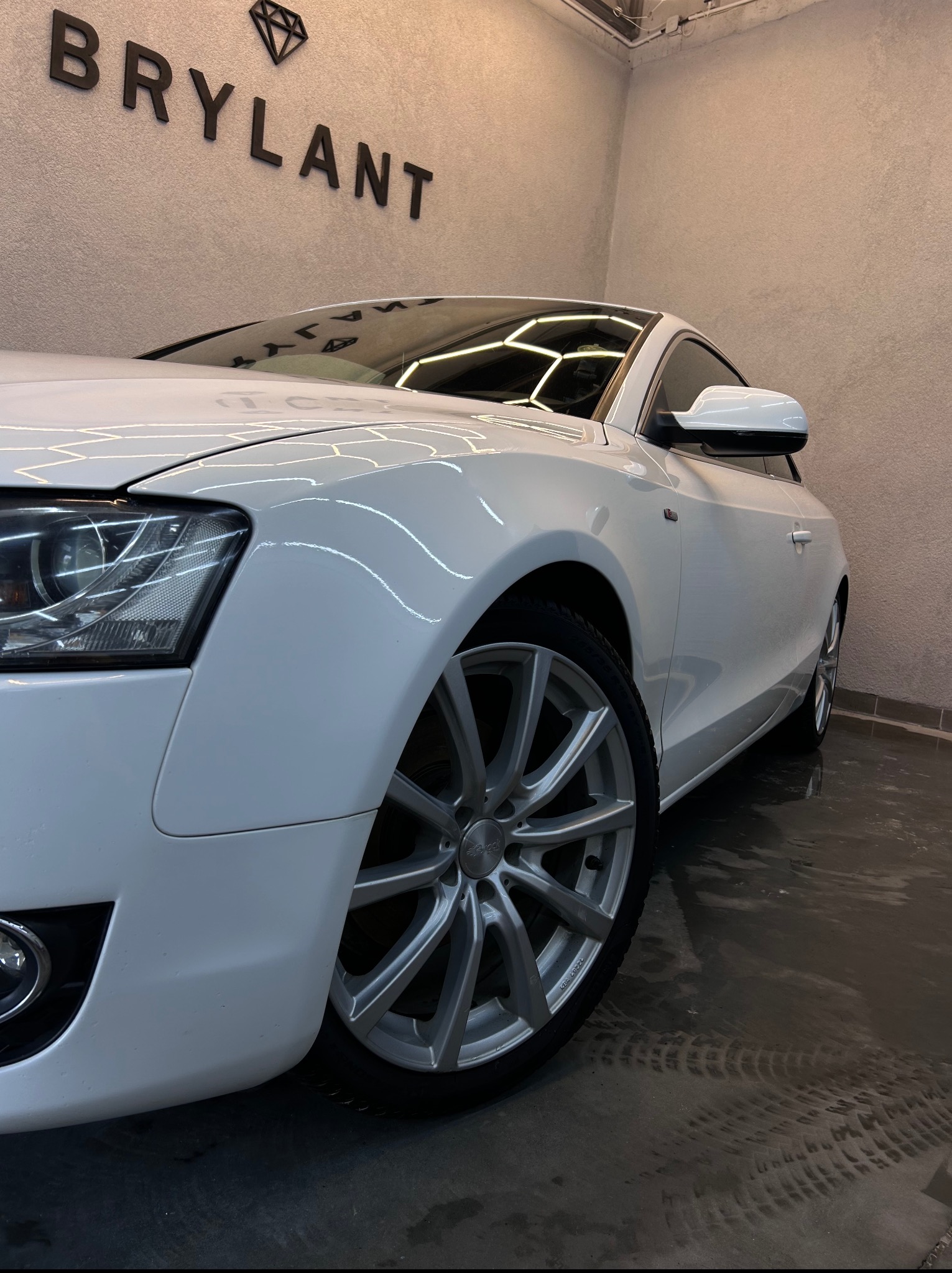 Białe Audi A5 po detailingu, z widocznym efektem hydrofobowym na lakierze i czystych felgach. Logo firmy detailingowej na ścianie w tle.