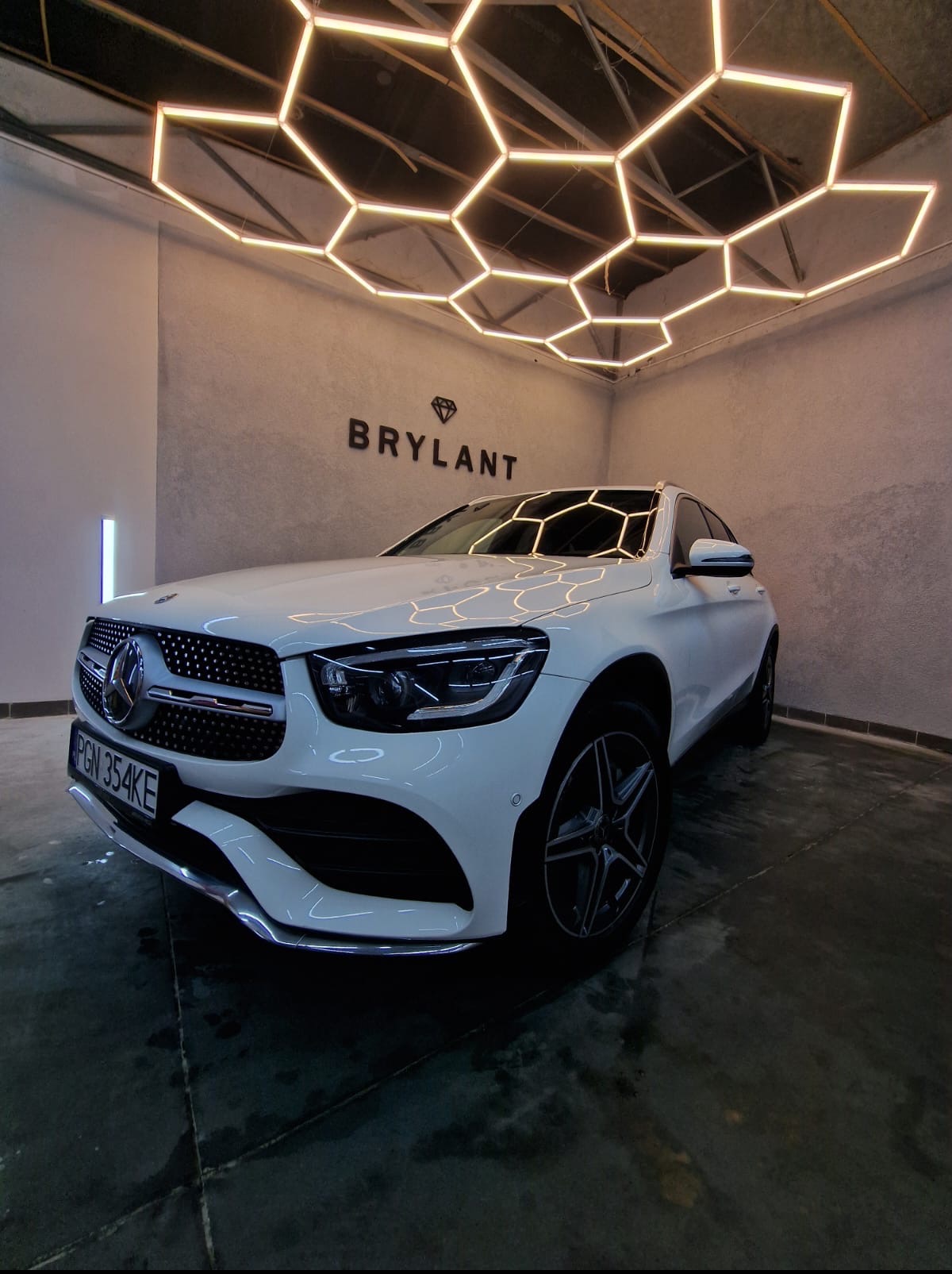 Biały Mercedes GLC po detailingu w studiu Brylant, oświetlony heksagonalnymi lampami. Błyszcząca karoseria, widoczny numer rejestracyjny. Profesjonalne oświetlenie.