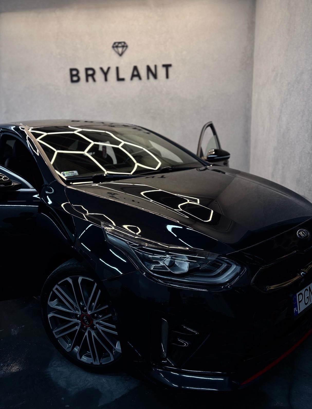 Błyszczący, czarny samochód Kia z otwartymi drzwiami, odbijający światła w kształcie plastra miodu. Logo 'Brylant' na ścianie w tle. Auto po detailingu.