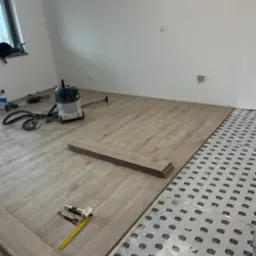 Układanie paneli podłogowych w jasnym odcieniu, widoczna warstwa izolacyjna, narzędzia pracy: miarka, nóż, młotek. W tle odkurzacz przemysłowy.