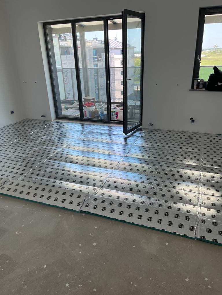Przygotowanie podłoża pod ogrzewanie podłogowe: rozłożone płyty izolacyjne z nadrukiem na surowym podkładzie. Duże okno balkonowe z otwartymi drzwiami, widok na budowę.