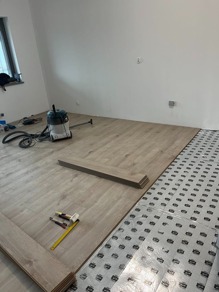 Układanie paneli podłogowych w jasnym odcieniu, widoczna warstwa izolacyjna, narzędzia pracy: miarka, nóż, młotek. W tle odkurzacz przemysłowy.