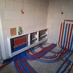 pompyhydraulika - Rozdzielacz ogrzewania podłogowego z rurami w kolorach czerwonym i niebieskim, zamontowany na ścianie z betonu komórkowego. Widoczna butelka i kartka z notatkami.