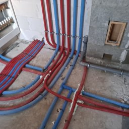 pompyhydraulika - Instalacja ogrzewania podłogowego: rury czerwone i niebieskie układane na podłodze, przymocowane do ściany. Widoczny rozdzielacz i fragment ściany z pustaków.