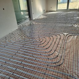 pompyhydraulika - Białe rury ogrzewania podłogowego ułożone w pętle na czerwonej siatce w jasnym pomieszczeniu z dużymi oknami. Etap instalacji przed wylaniem wylewki.