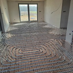 pompyhydraulika - Instalacja ogrzewania podłogowego w jasnym wnętrzu. Widoczne białe rury ułożone na czerwonej siatce, okno w tle.