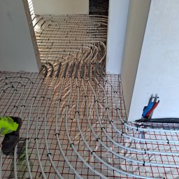 pompyhydraulika - Białe rury ogrzewania podłogowego, mocowane czarnymi uchwytami na folii z czerwoną siatką, prowadzone do ściany z wyprowadzonymi rurami instalacji.