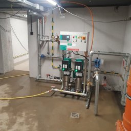 pompyhydraulika - Kompleksowa instalacja hydroforni w piwnicy budynku: dwa pionowe zbiorniki, panel sterowania, rury, zawory, manometry. Widoczna instalacja elektryczna i odpływ podłogowy.