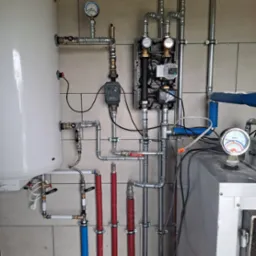 Złożona instalacja grzewcza z rurami, zaworami i manometrami na beżowej ścianie z płytek. Elementy w kolorze srebrnym, czerwonym i niebieskim. Widoczny bojler i piec.