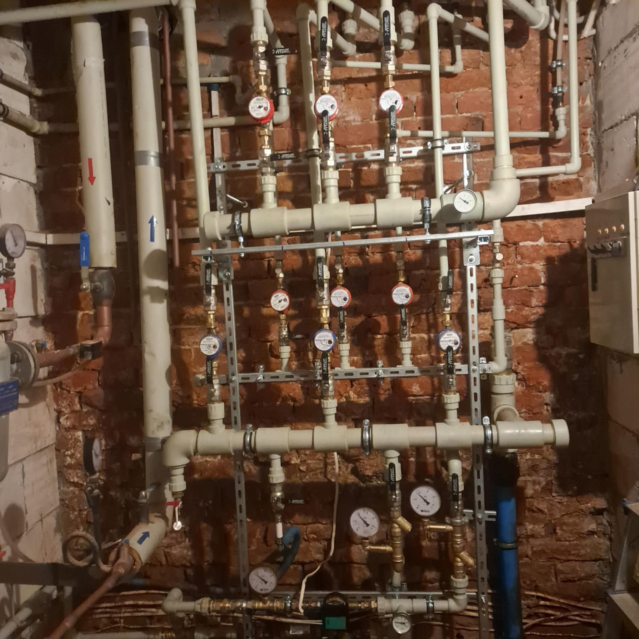 Rozbudowana instalacja grzewcza z rurami, zaworami i manometrami na tle ceglanej ściany. Widoczne elementy mocujące i fragment skrzynki elektrycznej.