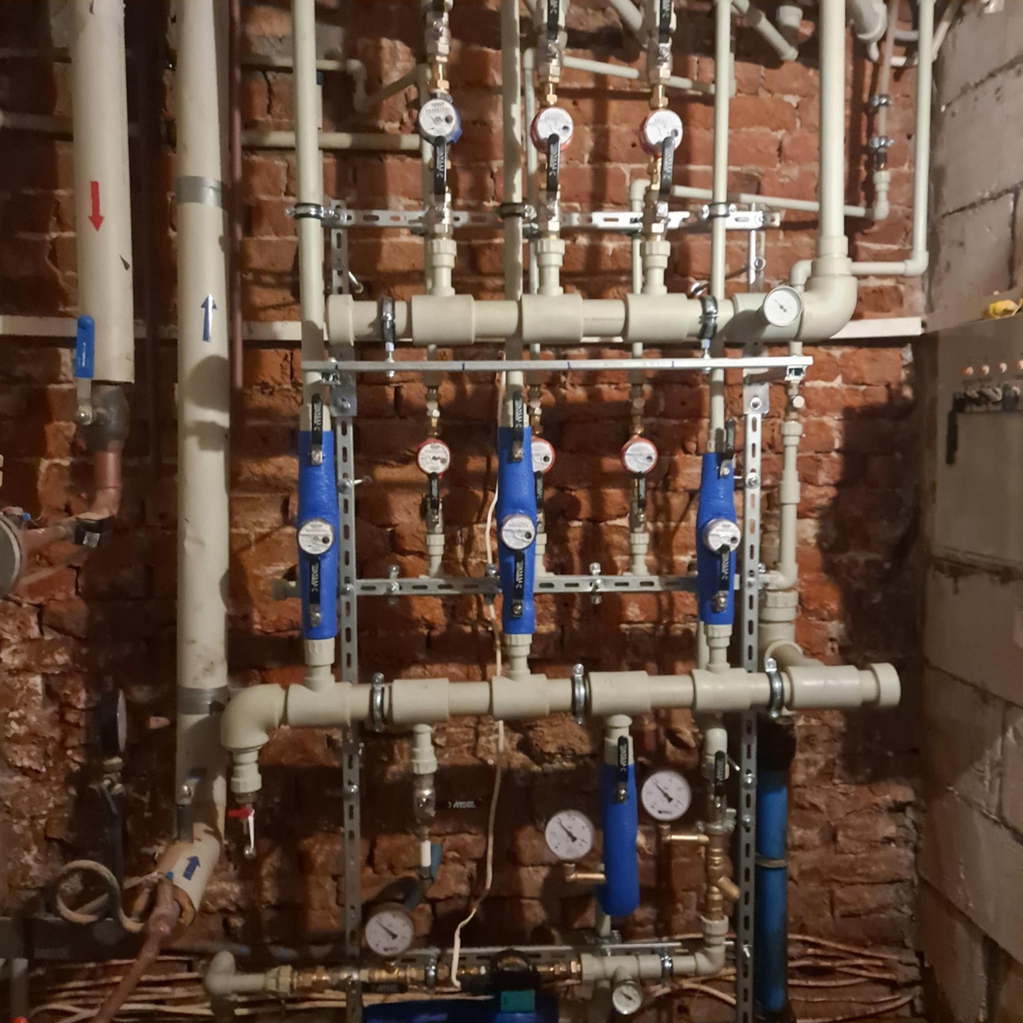 Złożona instalacja hydrauliczna z rurami, zaworami i manometrami na tle ceglanej ściany. Widoczne elementy mocujące i przewody.