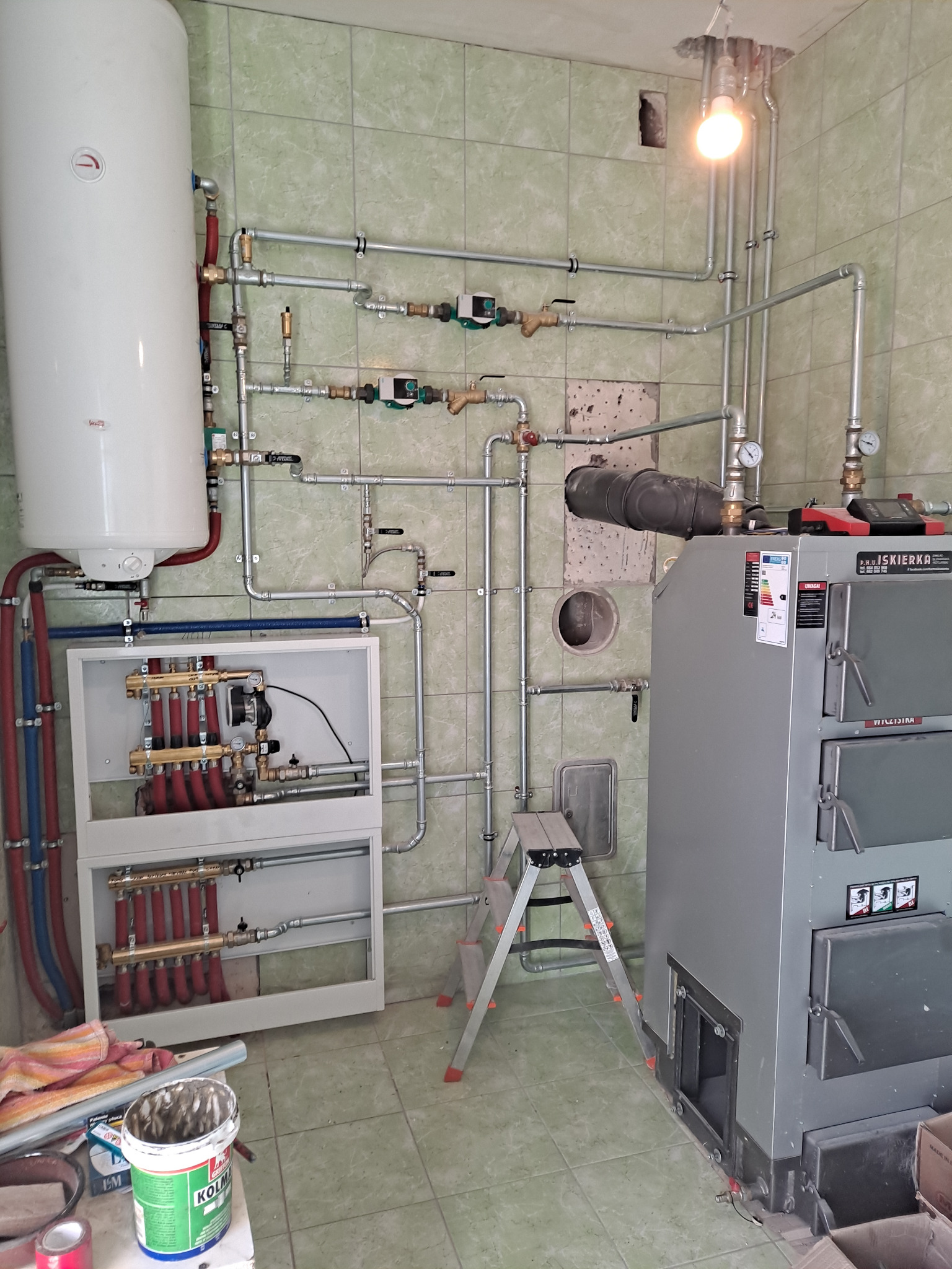 Kompleksowa instalacja grzewcza z piecem węglowym i zasobnikiem wody, widoczna na tle ściany z zielonych płytek. Rury, zawory i rozdzielacz tworzą skomplikowany system.