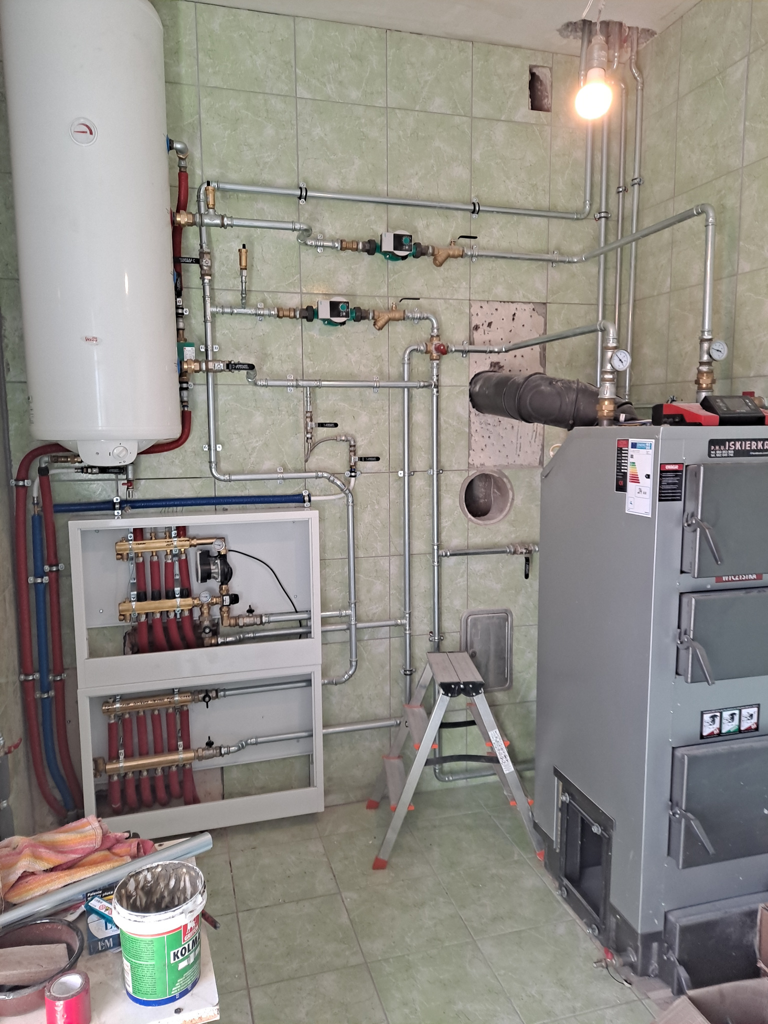 Kotłownia z piecem, bojlerem i rozdzielaczem, widoczne rury instalacji grzewczej na tle zielonych płytek. U dołu drabina i narzędzia.