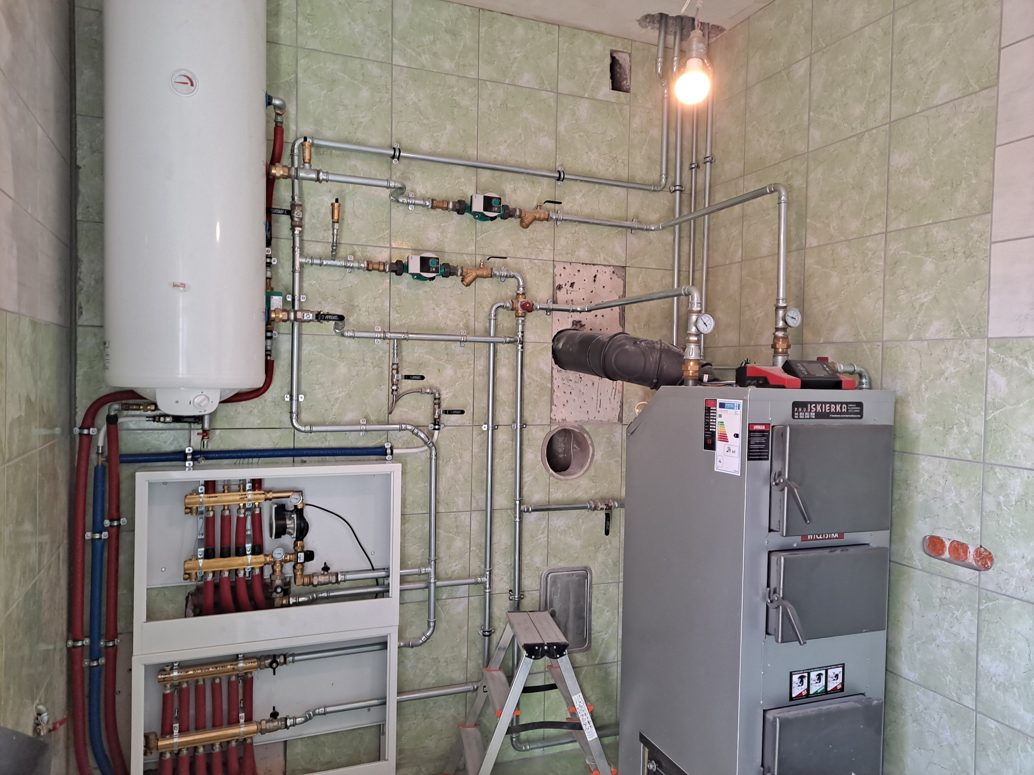 Złożona instalacja grzewcza z piecem, zbiornikiem i rozdzielaczem, widoczna drabinka monterska. Szare płytki na ścianach. Ujęcie z dołu, widoczna żarówka.