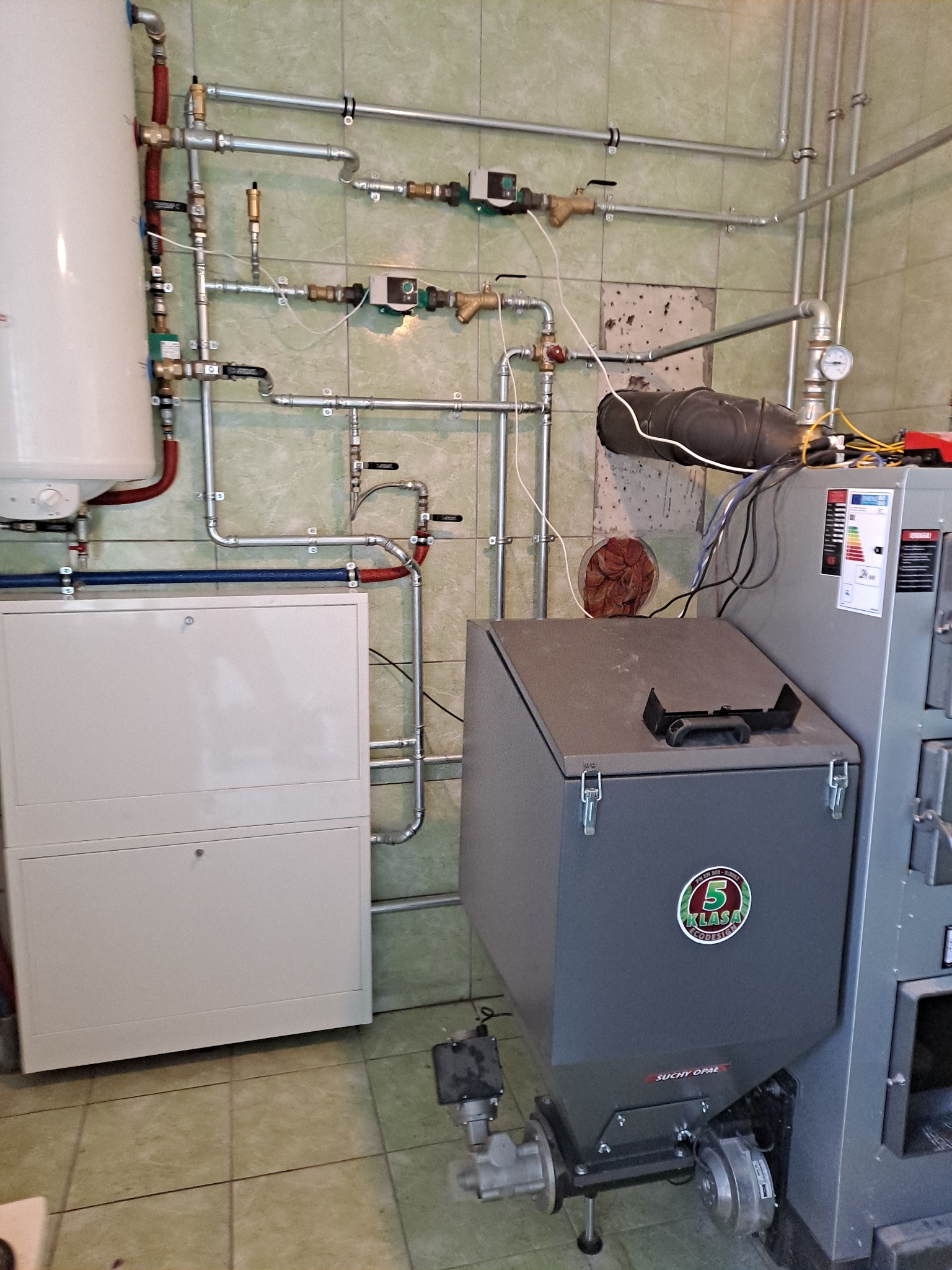 Złożona instalacja grzewcza z kotłem na ekogroszek, zasobnikiem ciepłej wody i rozbudowaną siecią rur miedzianych. Widoczne elementy sterujące i zabezpieczające.