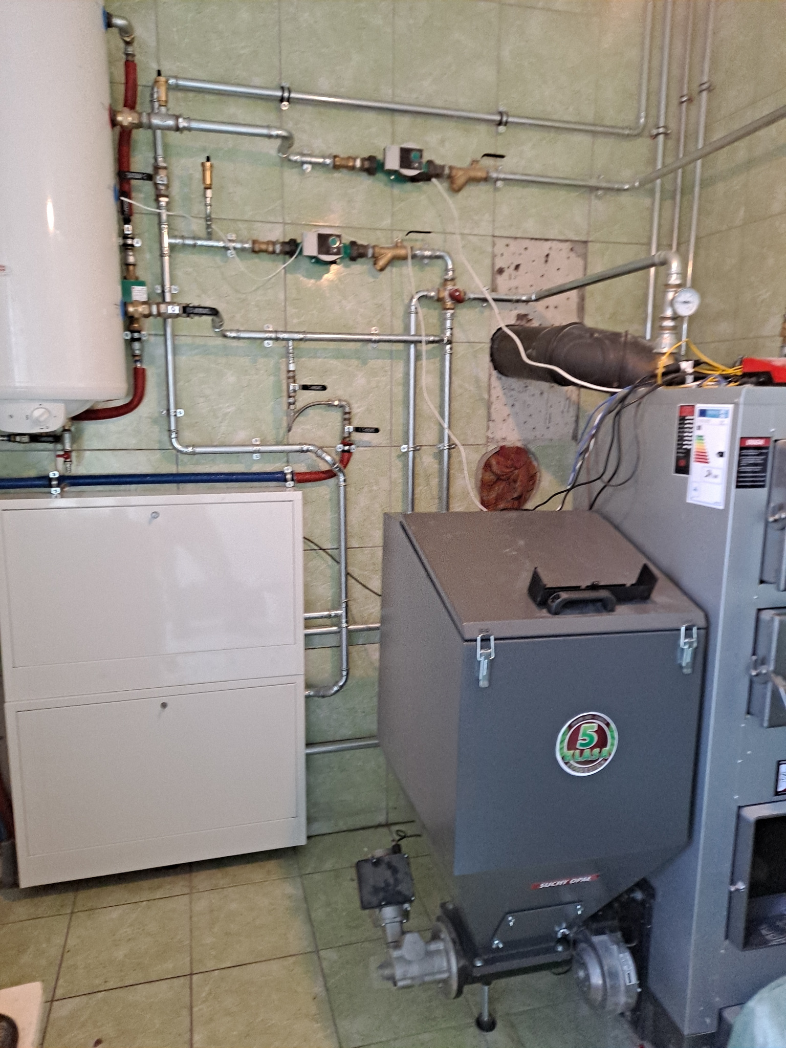Kompleksowa instalacja grzewcza z kotłem na paliwo stałe, rurami i zbiornikiem, widoczna na tle kafelkowanej ściany. Elementy instalacji starannie połączone.