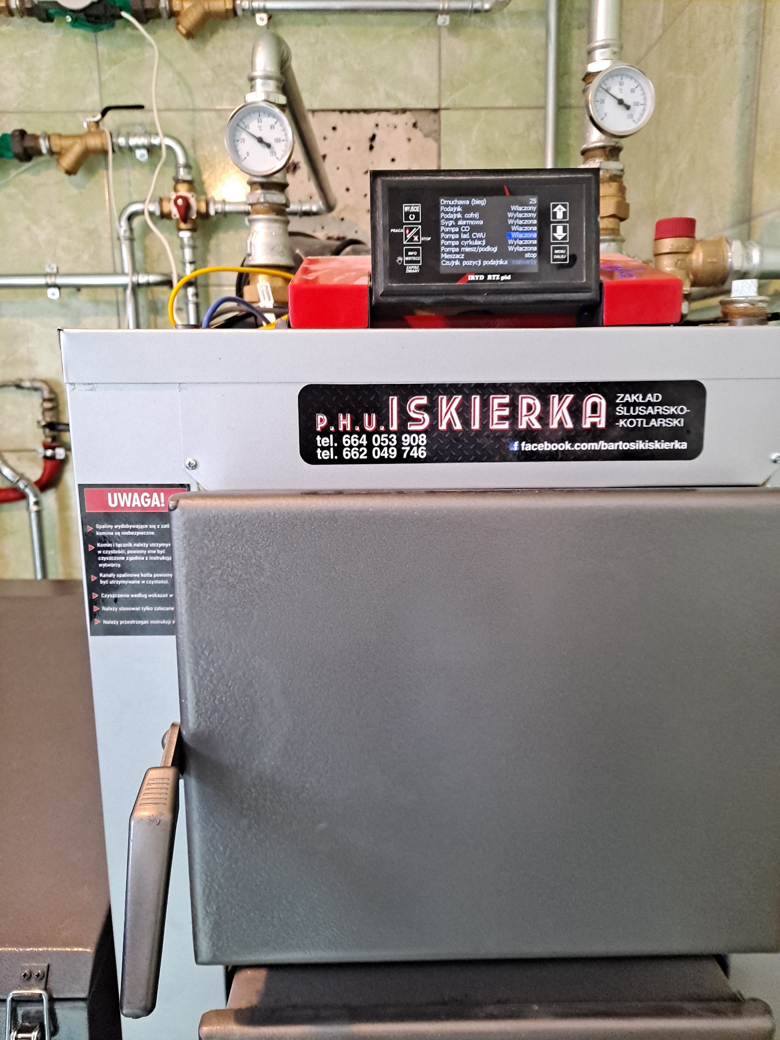 Kocioł grzewczy z panelem sterowania, widoczne rury instalacji i manometry. Na kotle naklejka z danymi firmy P.H.U. Iskierka.