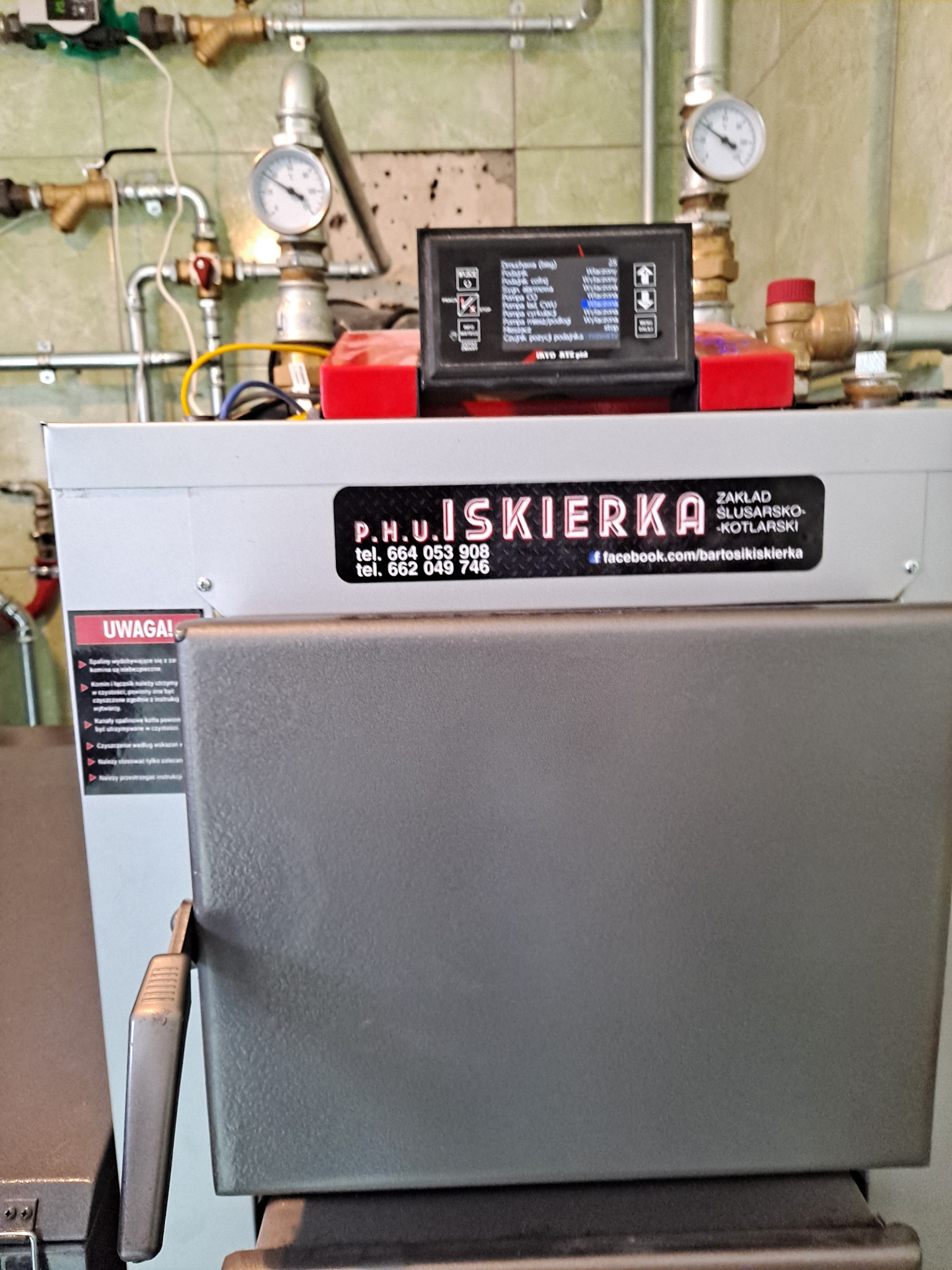 Szary piec CO z panelem sterowania, widoczne rury instalacji grzewczej i manometry. Na piecu naklejka z danymi firmy P.H.U. Iskierka.