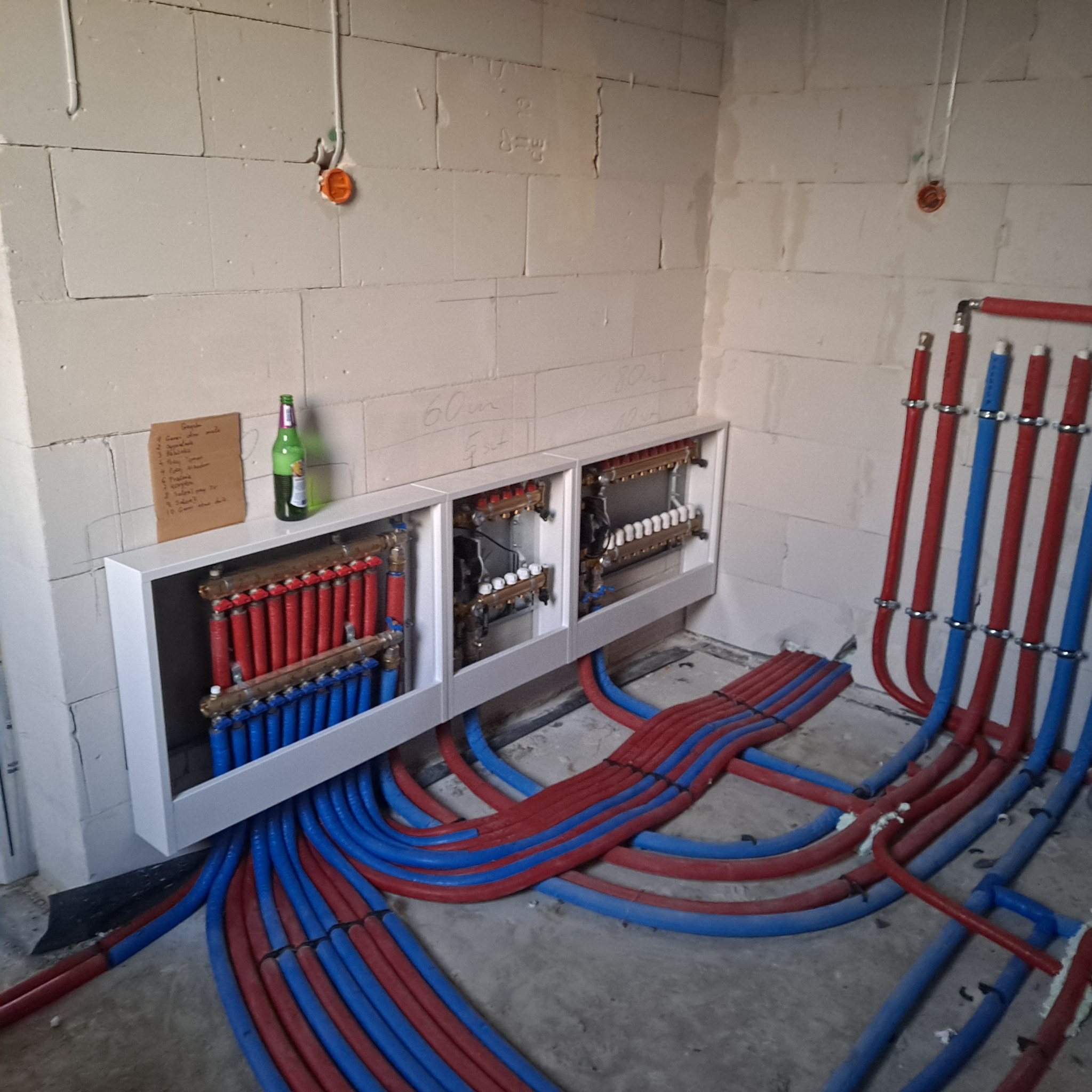 Rozdzielacz ogrzewania podłogowego z rurami w kolorach czerwonym i niebieskim, zamontowany na ścianie z betonu komórkowego. Widoczna butelka i kartka z notatkami.