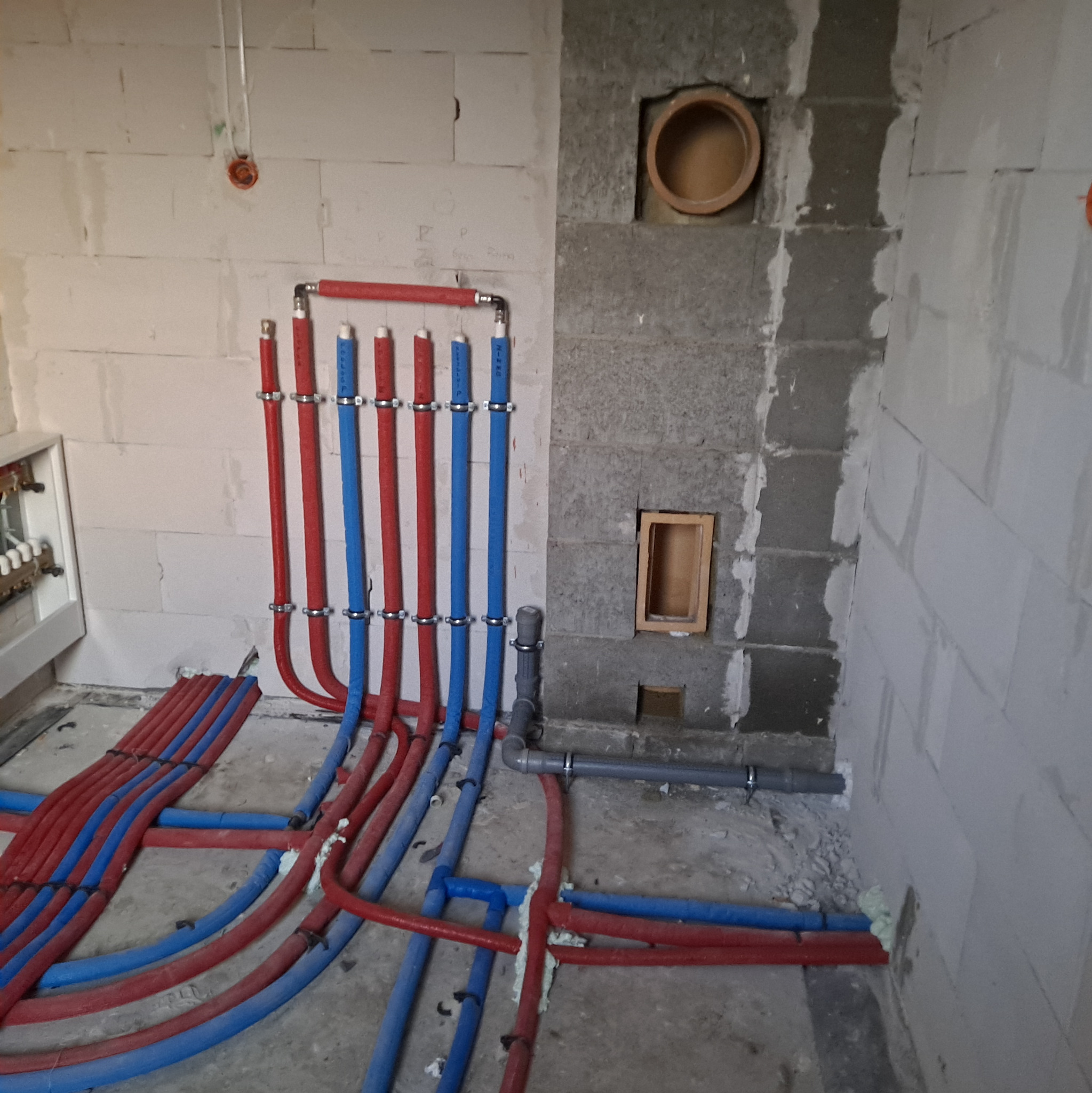 Instalacja wodna w surowym wnętrzu: pionowe rury czerwone i niebieskie, mocowane do ściany z betonu komórkowego, poziome rury na podłodze, otwory w ścianie.