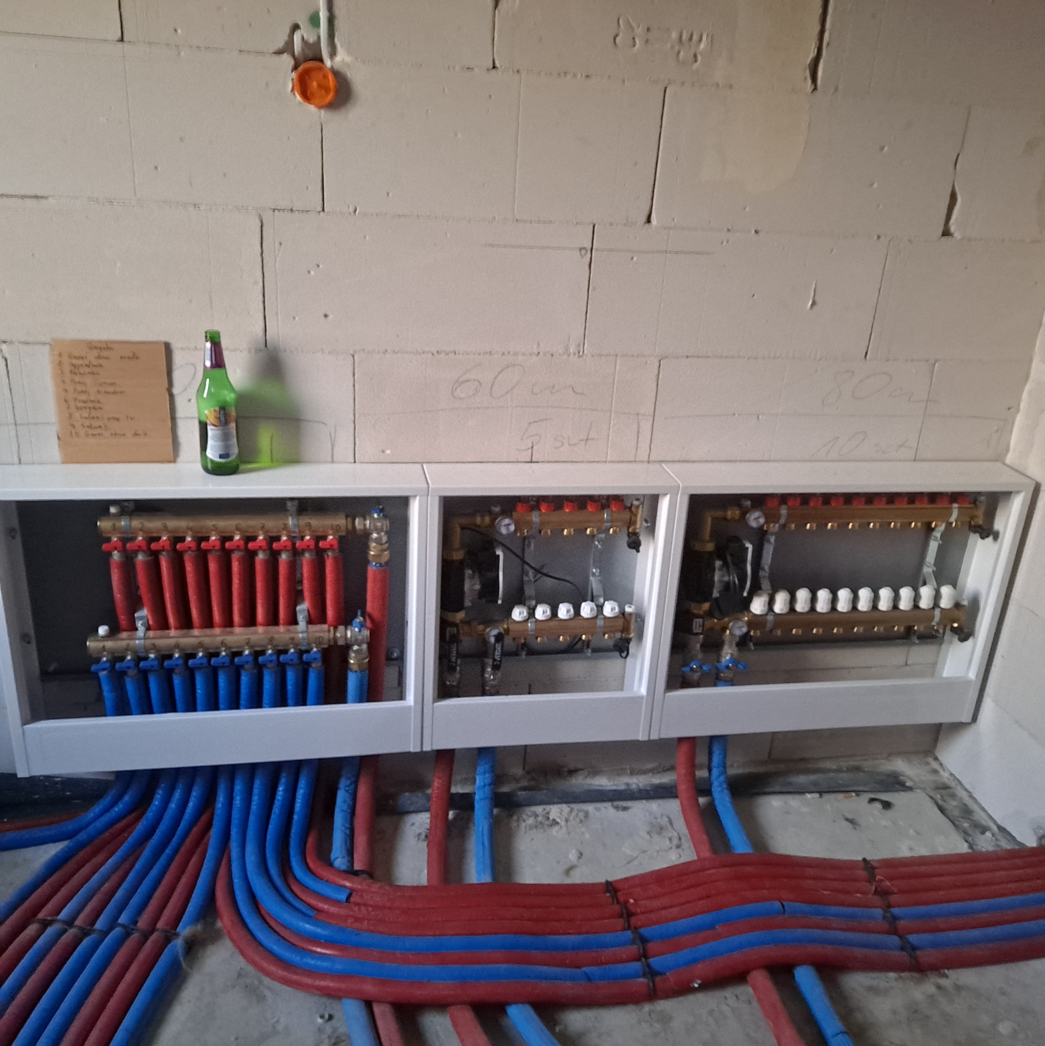 Rozdzielacz ogrzewania podłogowego w białej obudowie, z czerwonymi i niebieskimi rurami. Na ścianie notatka i butelka. Widoczne napisy na ścianie.