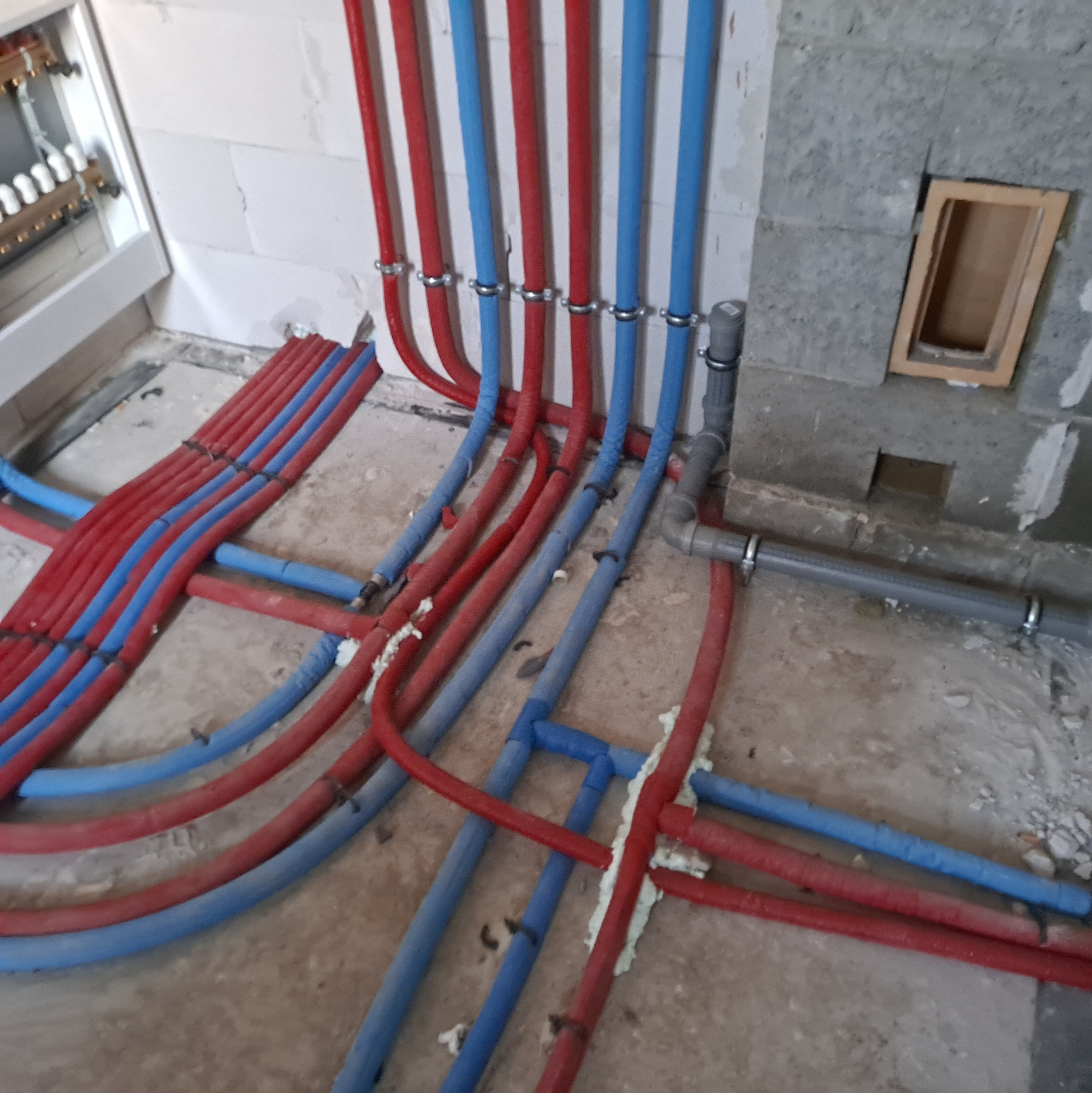 Instalacja ogrzewania podłogowego: rury czerwone i niebieskie układane na podłodze, przymocowane do ściany. Widoczny rozdzielacz i fragment ściany z pustaków.